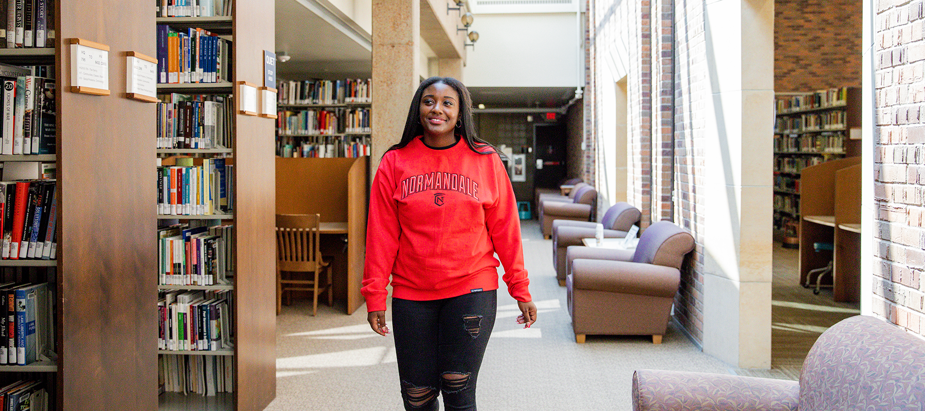 Smiling student in a red Normandale sweatshirt walks through the library.