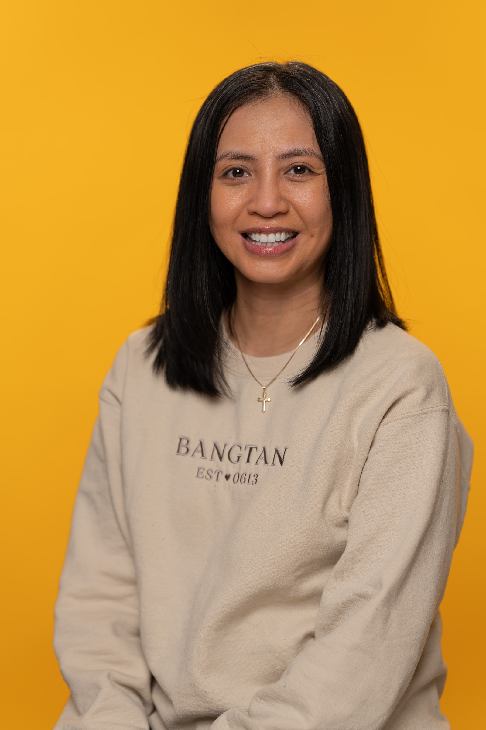 Loraine Navales - Psychology Student of the Month,Photo of Loraine Navales, a woman with brown hair that goes past her shoulders. She is sitting in front of an orange background with a cream sweatshirt and wearing a cross.