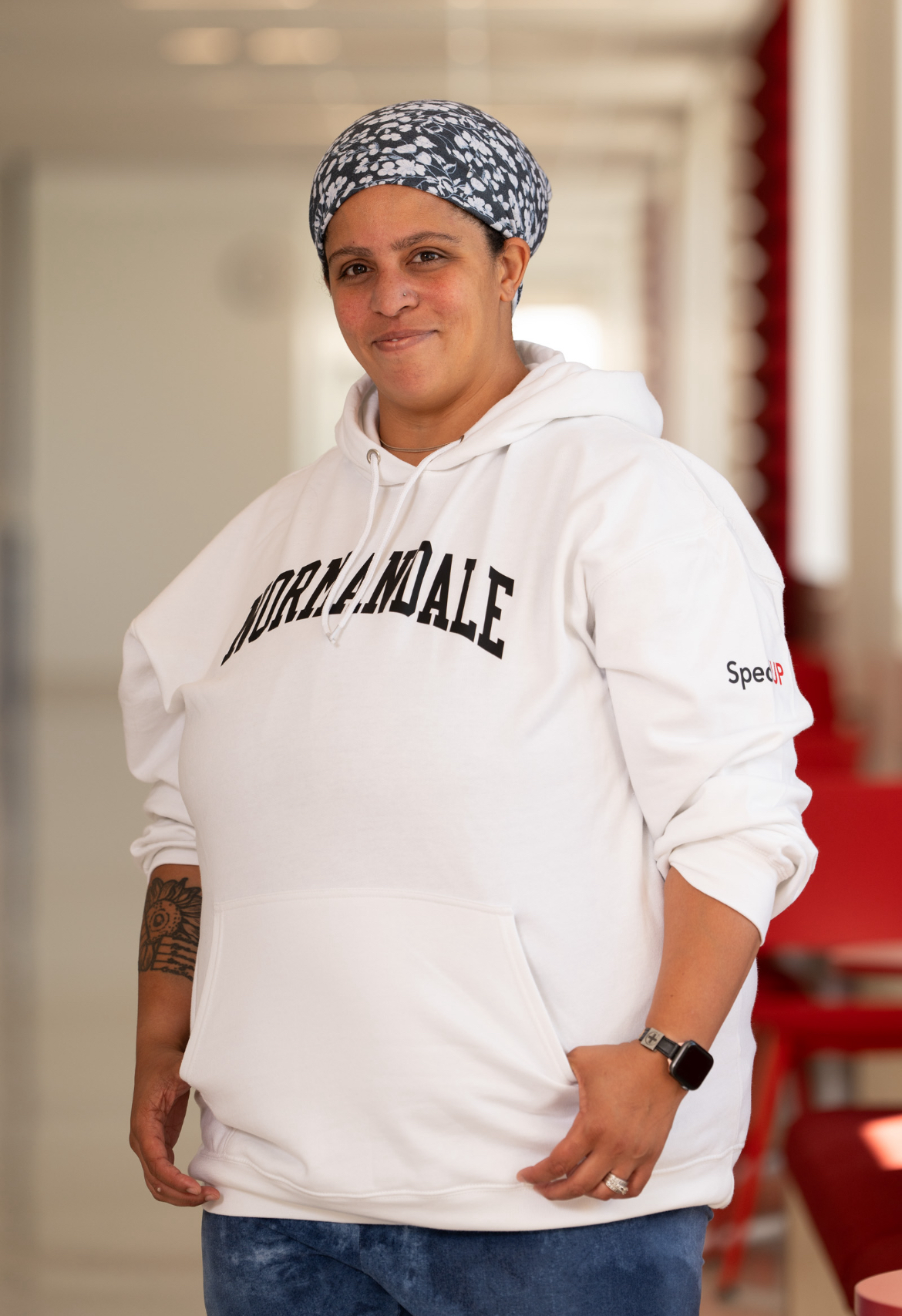 Individual with white sweatshirt that says Normandale. Black and white head covering. Watch and ring on one hand and tattoo on other arm. Standing in hallway of Normandale. Thumbs are in pockets of sweatshirt.